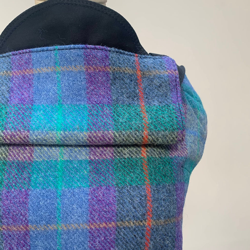 A person wearing a Harris Tweed Aurora Integra Baby Carrier in a plaid pattern with colors blue, green, and purple.