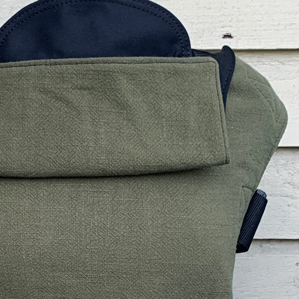 A khaki textured linen baby carrier with black cotton straps, Size 1, displayed against a white wooden background.